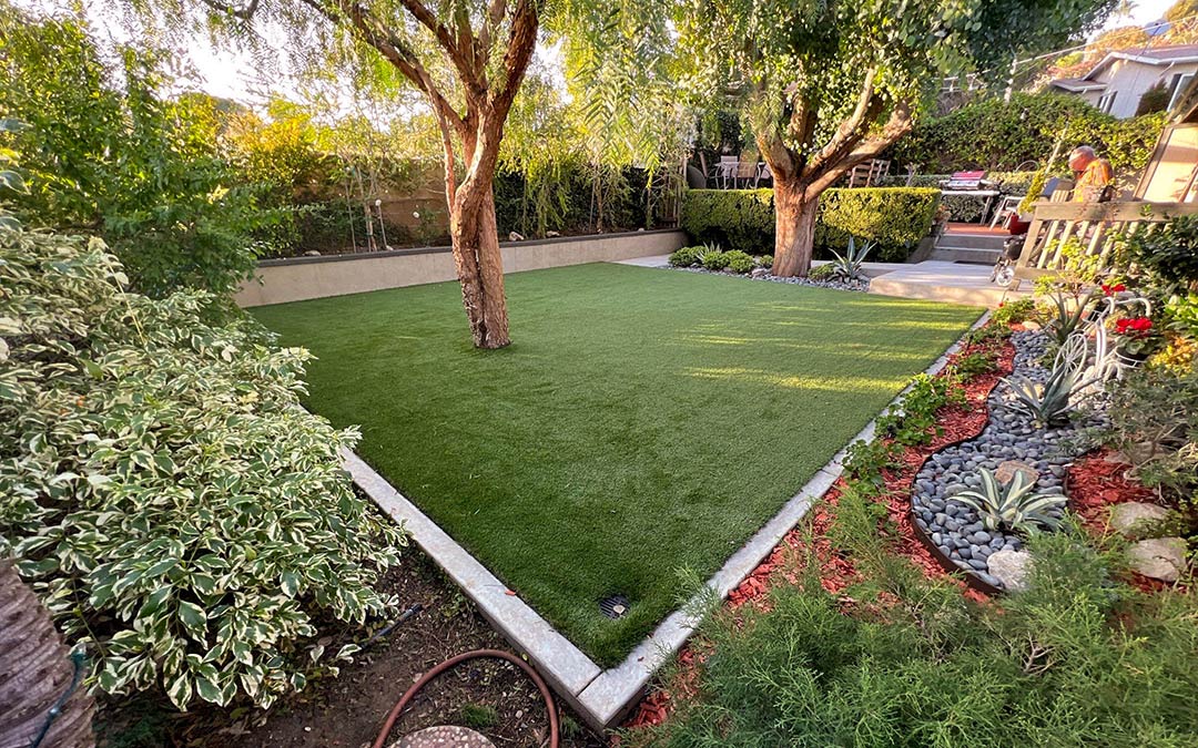 Image of a backyard with artificial grass and trees and a perimeter of blushes and plants